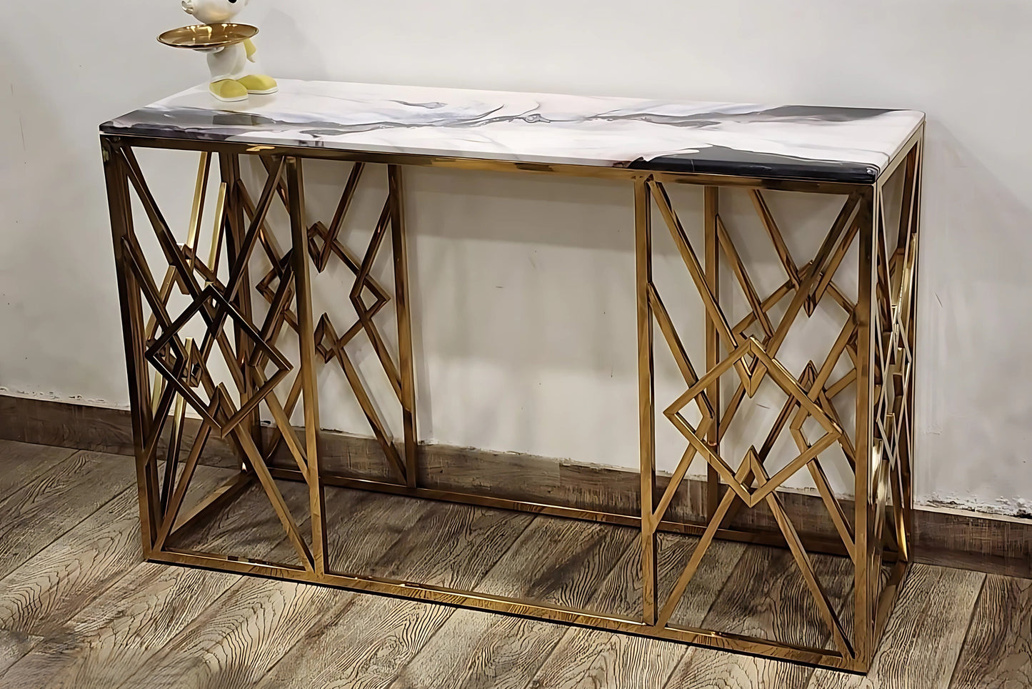 A white marble console table with gold looped legs, placed against a wall on a wooden floor with a decorative item on top.