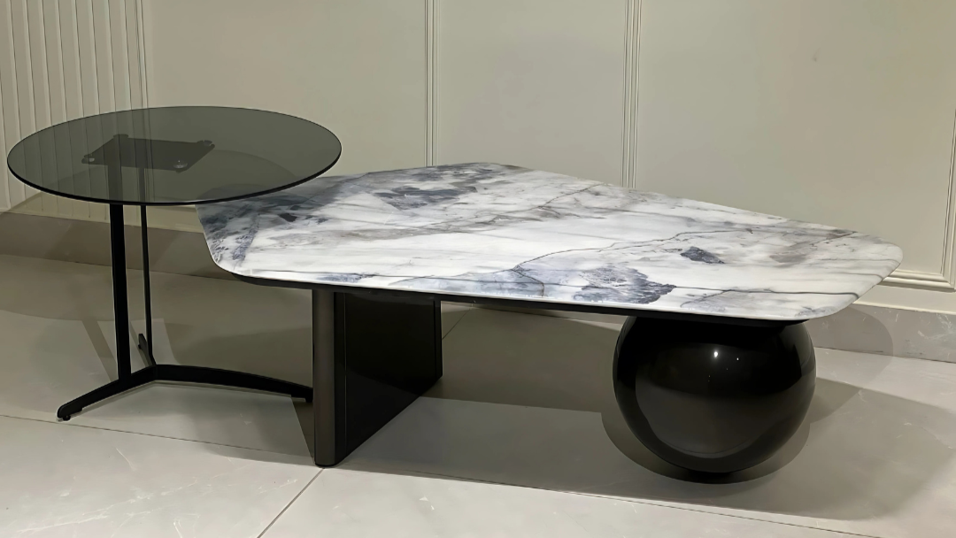 Modern asymmetrical marble and glass center coffee table with sculptural black base and curved detailing in a minimalist interior.