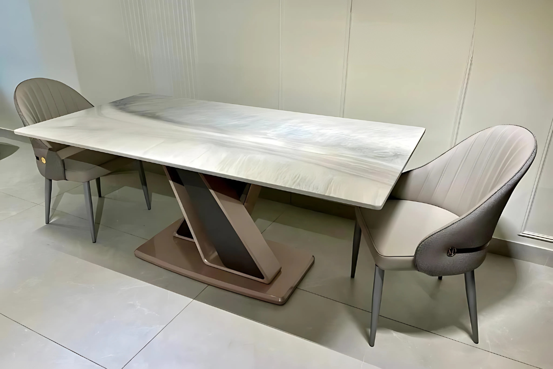Luxury dining table with a grey marble-like tabletop and a bronze metal pedestal base, flanked by two modern chairs with beige and brown upholstery.